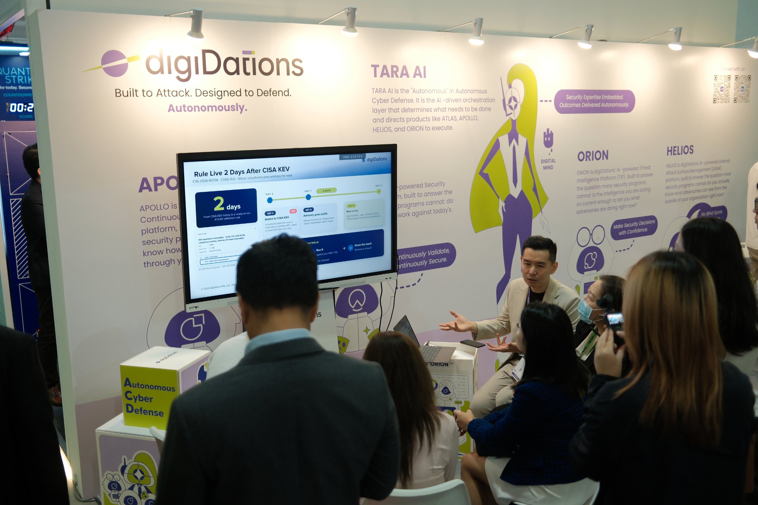 A digiDations team member talks with a group of visitors seated around him at Booth D-02, in front of a display and the TARA AI, ORION and HELIOS wall graphics.