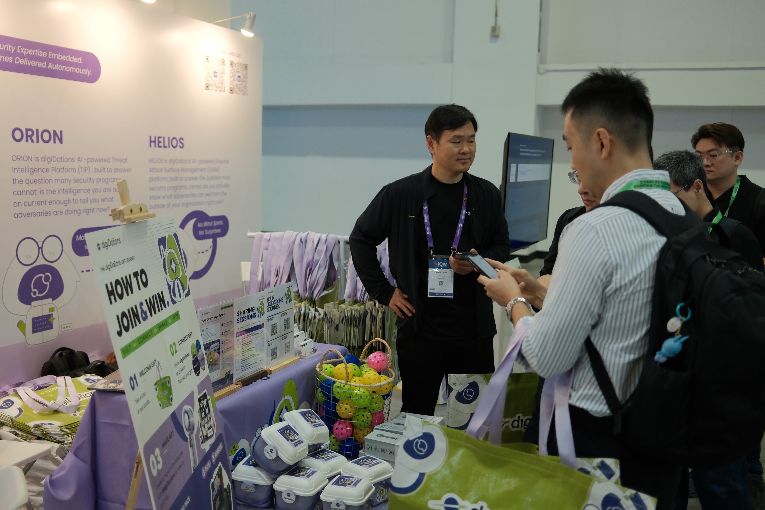 Visitors at the Booth D-02 table beside the How to Join and Win gift journey sign, with digiDations tote bags and gift boxes.